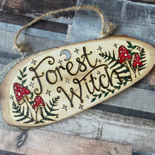 Load image into Gallery viewer, Wooden oval sign with live bark edges featuring the words Forest Witch and a design of red and white mushrooms, ferns, crescent moon and stars, burned using the art of Pyrography and painted. Has a string for hanging