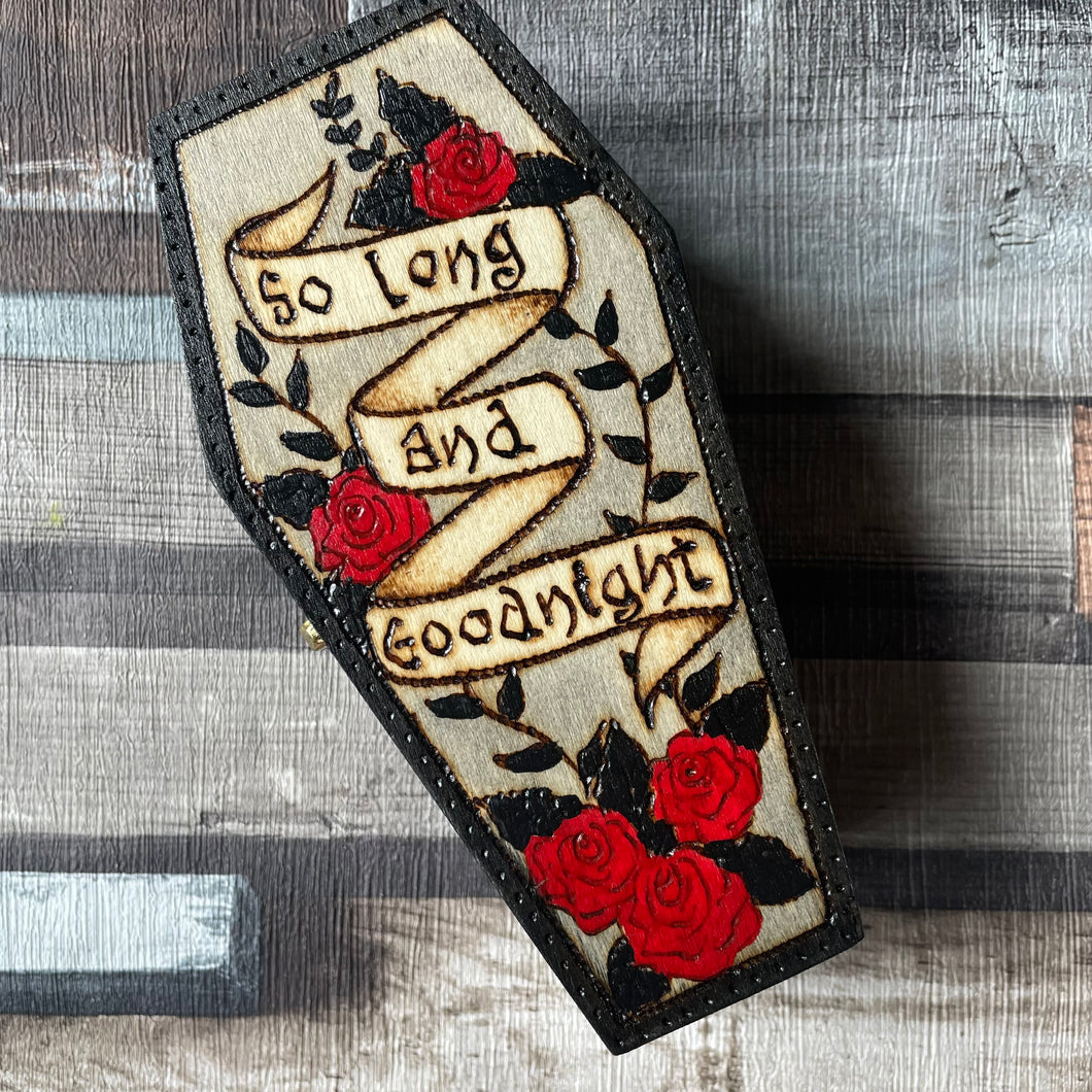 Wooden box in a coffin shape, with the words So long and Goodnight on a banner, red roses and black foliage 