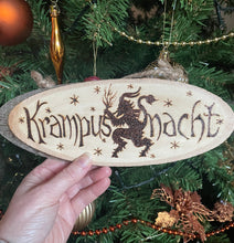 Load image into Gallery viewer, Oval Wood slice featuring a Krampus image and the words “Krampusnacht” burned onto it. It has hooks and string for hanging 